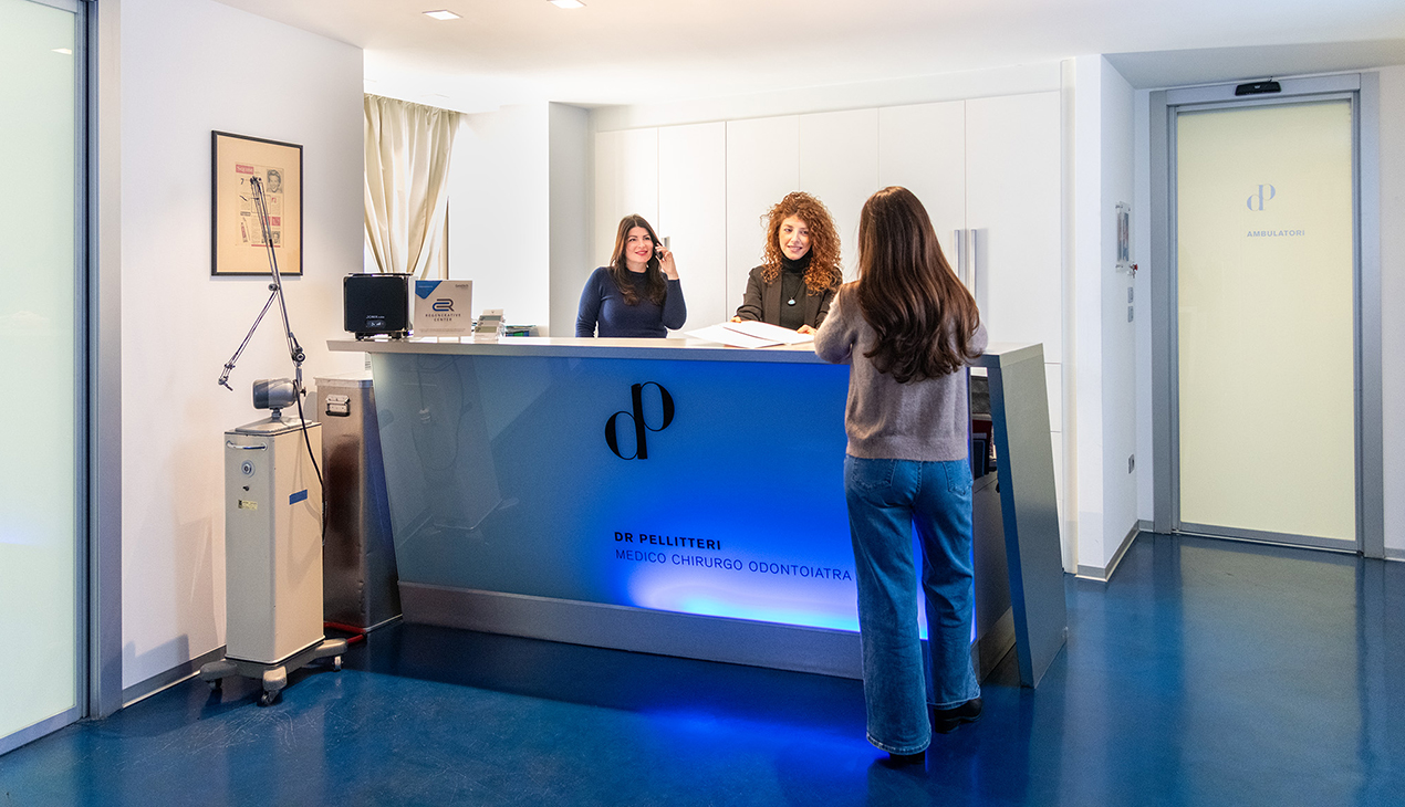 Due assistenti accolgono una paziente alla reception illuminata di blu dello studio dentistico Pellitteri a Bolzano